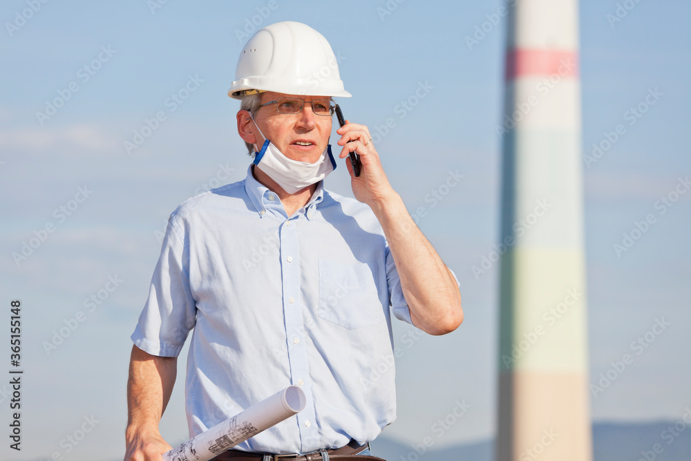 Engineer or architect with face mask and blueprint in his hand on a construction site talking on cell phone Engineer or architect with face mask and blueprint in his hand on a construction site talking on cell phone