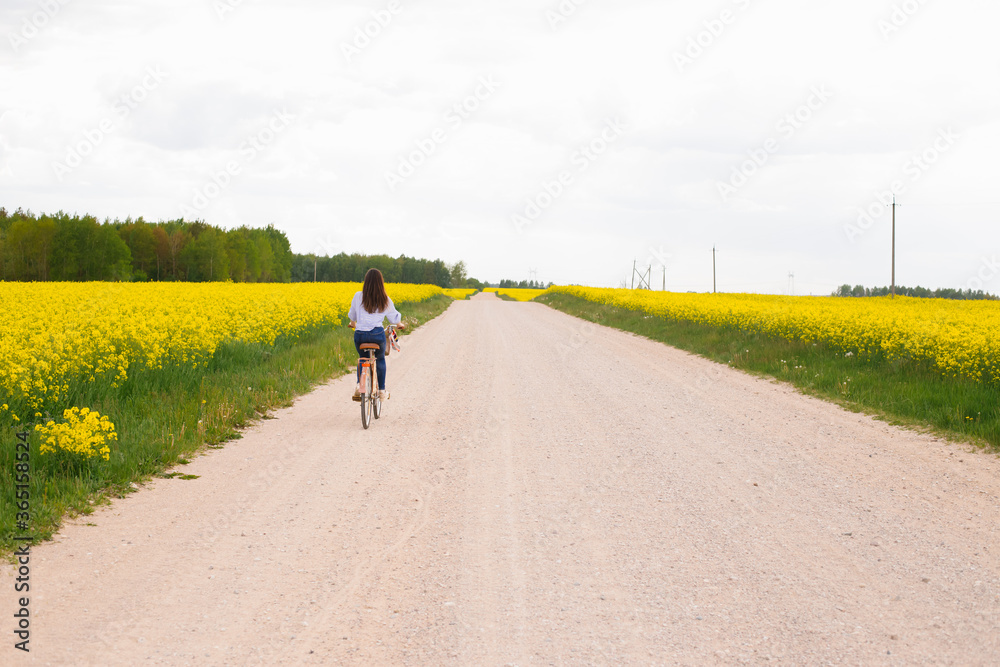 Fototapeta premium Young woman rides a bicycle on a road between a blooming rapeseed field. Yellow flowering rapeseed field. Bright spring bloom of rapeseed
