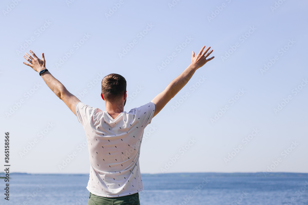 A young man stands facing the sea with his hands up. Summer freedom. Summer vacation by the sea