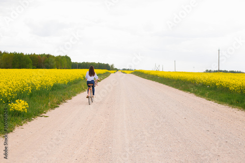 Young woman rides a bicycle on a road between a blooming rapeseed field. Yellow flowering rapeseed field. Bright spring bloom of rapeseed