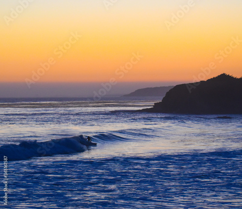 Brilliant orange and purple sunset reflection on big Ocean blue wave that surfer is surfing