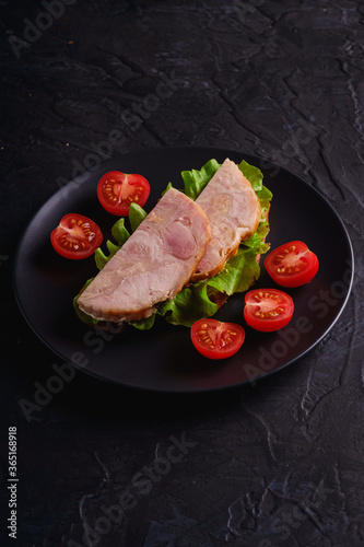Sandwich with turkey ham meat, green salad and fresh cherry tomatoes slices on black plate, dark textured background, angle view