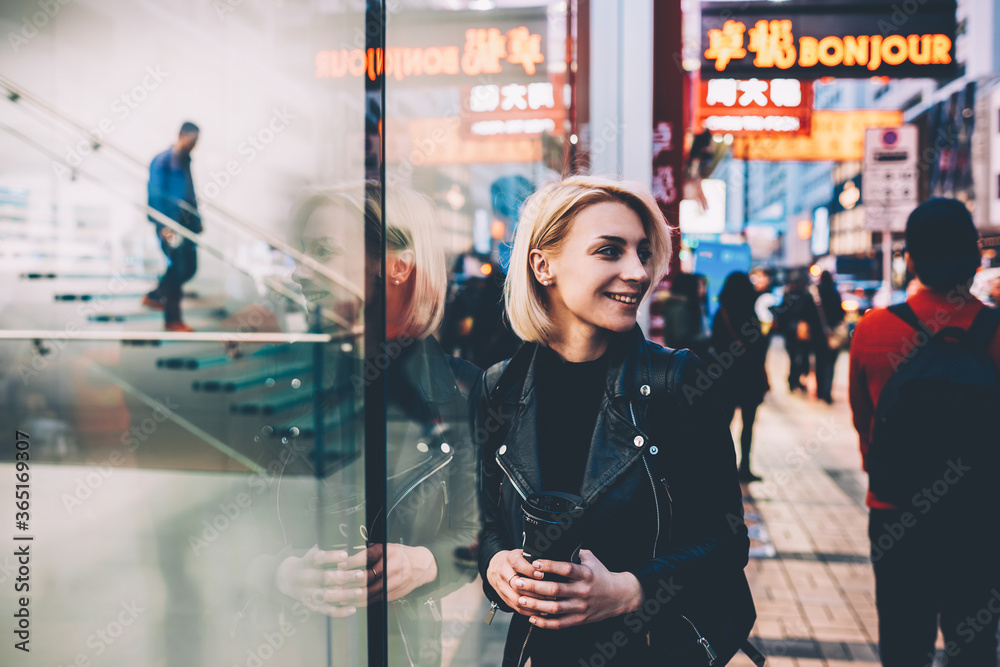 Cheerful blonde young woman dressed in casual leather jacket laughing while strolling on streets with tasty beverage in hand.Positive hipster girl laughing during walking outdoors with coffee to go