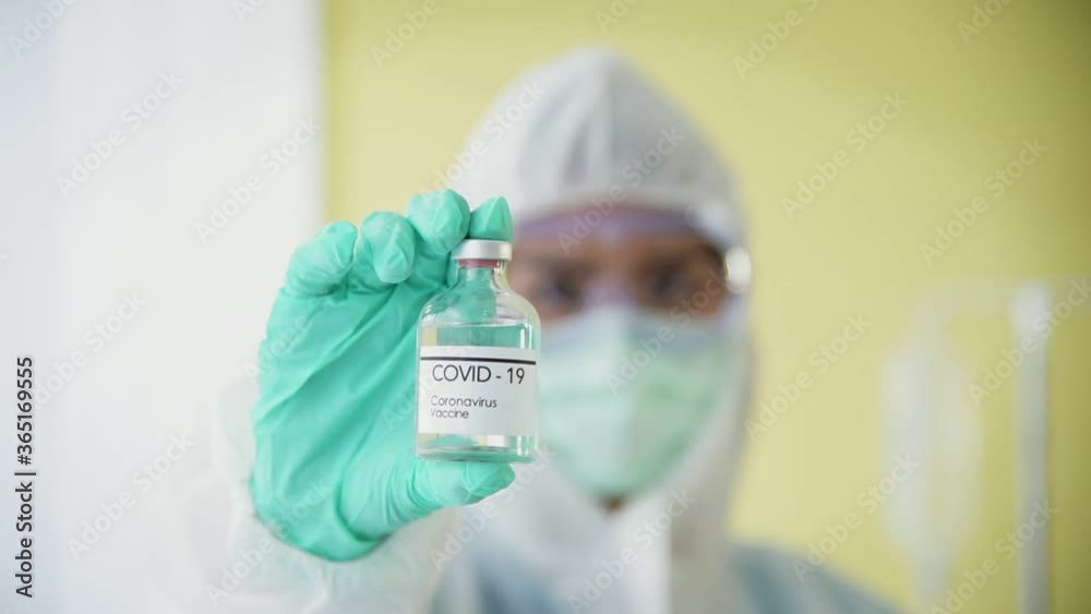 Professional doctor in sterile personal protective equipments holding liquid vaccine in bottle in white clean room background Professional doctor in sterile personal protective equipments holding liquid vaccine in bottle in white clean room background
