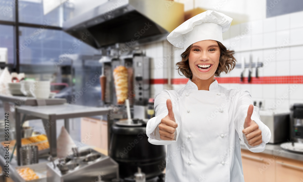 cooking, gesture and people concept - happy smiling female chef in toque showing thumbs up over restaurant or kebab shop kitchen background