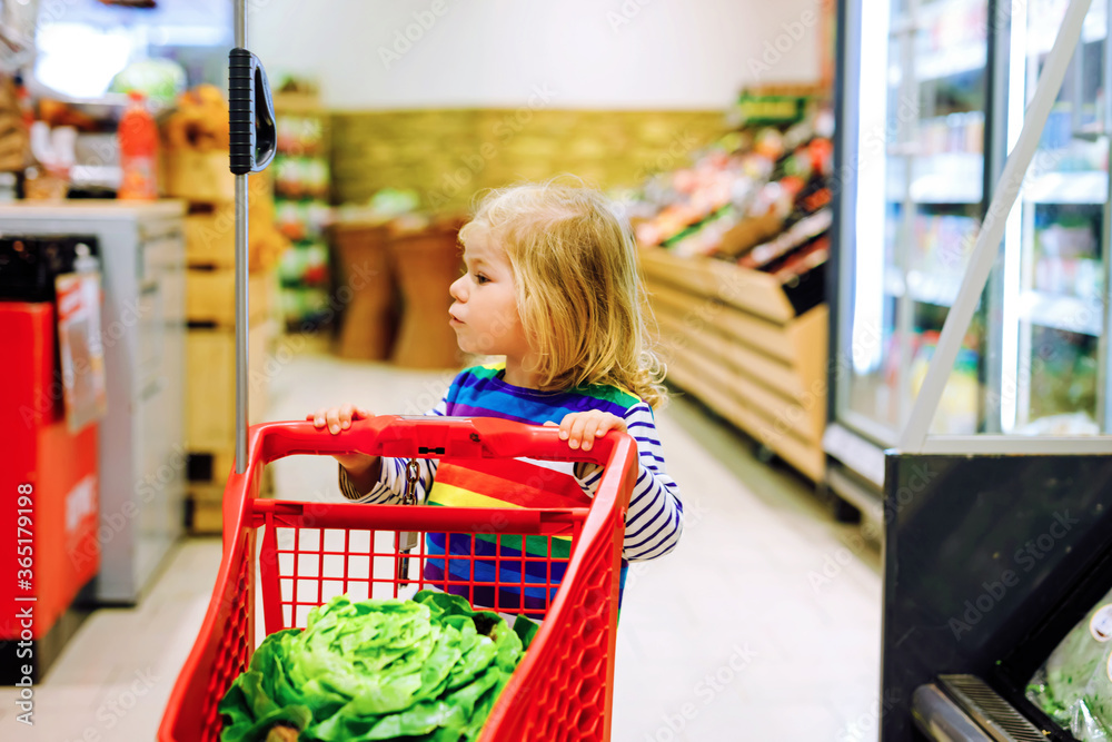 Cute todler girl pushing shopping cart in supermarket. Little child buying fruits. Kid grocery shopping. Adorable baby kid with trolley choosing fresh vegetables in local store.