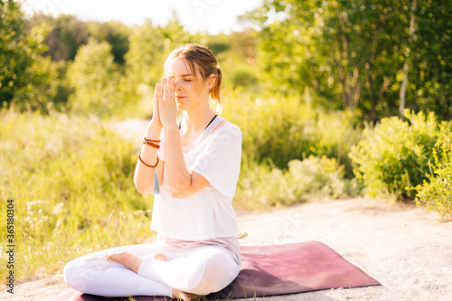 Young woman is meditating in lotus position with closed eyes sitting on yoga mat on grass. Female yoga instructor doing namaste yoga pose background of sunray outdoor on park outside the city.