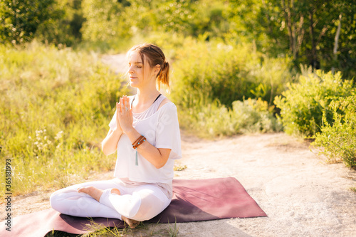 Relaxed young woman is meditating in lotus position with closed eyes sitting on yoga mat. Female yoga instructor doing namaste pose. Lady enjoying meditation. Girl prayer meditating in Namaste mudra.
