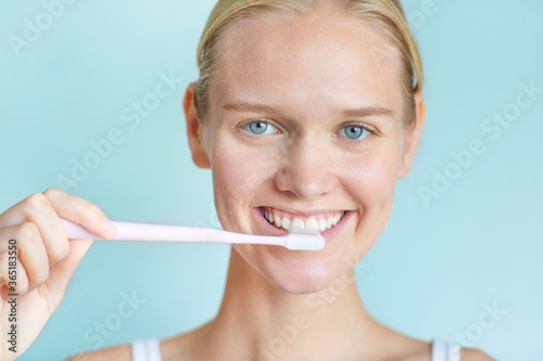 Happy young woman brushing her teeth white while looking at the camera smiling with beautiful eyes. Isolated against a blue background.