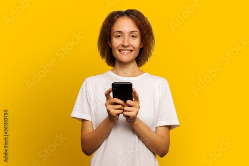 Portrait of a woman with curly hair in a white tank top holding a smartphone and smiling while looking at the camera on an isolated yellow background. Woman writes sms. Good news