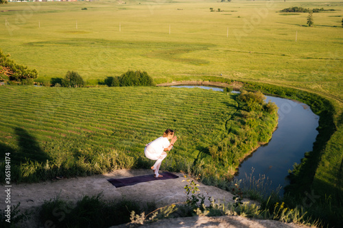 Young relaxed woman practicing yoga, standing in Eagle exercise, Garudasana pose on top of cliff background of river below and green grass in evening during sunset. 