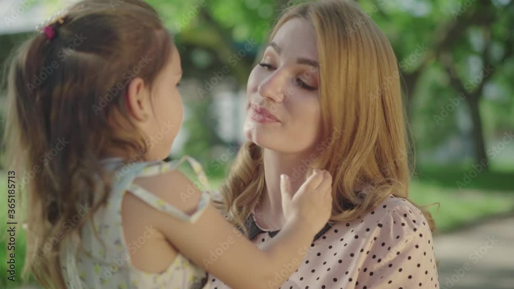 Close-up portrait of beautiful young Caucasian mother kissing pretty daughter in sunny park. Smiling blond woman spending happy summer day outdoors with child. Motherhood, happiness, joy. Close-up portrait of beautiful young Caucasian mother kissing pretty daughter in sunny park. Smiling blond woman spending happy summer day outdoors with child. Motherhood, happiness, joy.