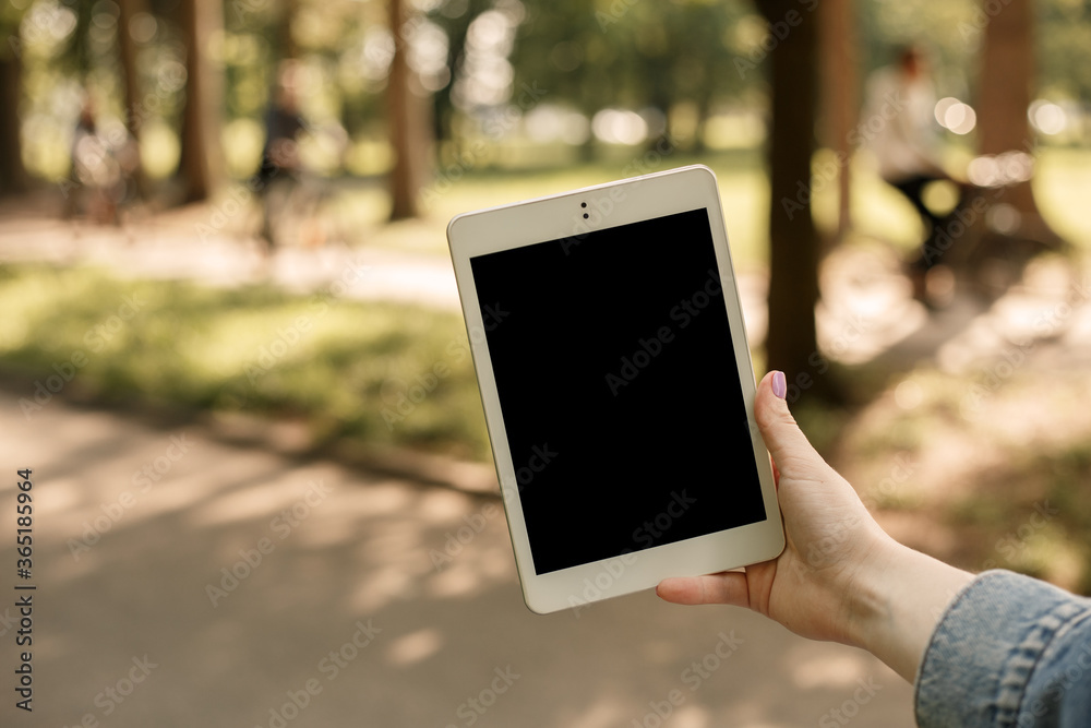 Close up shot of female hand holding white tablet pc in the city park in sunny weather. Mock up, copy space with people walking by on the blur background. 5G internet, network concept.