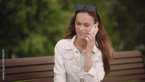 Pretty brunette woman is talking on cell phone while sitting on a bench in the park. Outdoors portrait