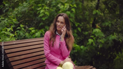 beautiful young woman is wearing bright clothes sitting on a bench in the park and talking on a smartphone