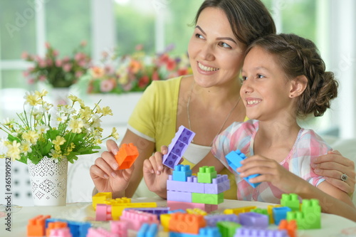 Little daughter and mother playing with colorful plastic blocks at home