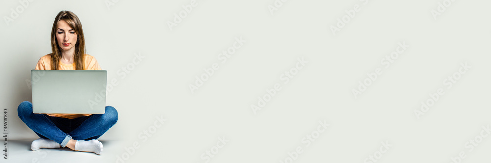 Naklejka premium Beautiful young girl sitting in lotus position, with a laptop on her knees on a light background. Banner.
