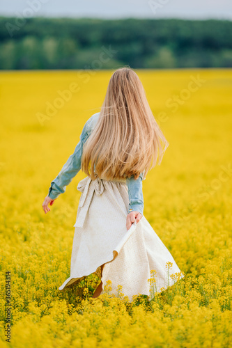 young woman in a dress in a yellow field, canola, walks her back through the flowers. blonde with long hair in nature, outside the house, walk in the field