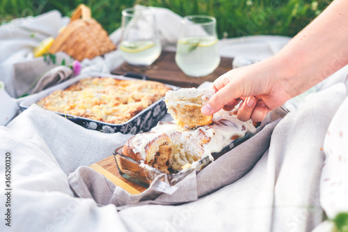 Woman takes homemade cinnabon by hand on white blanket outdoors. Picnic composition with two glasses of lemonade, peach pie on green grass on white blanket outdoors. Summer vacation concept.