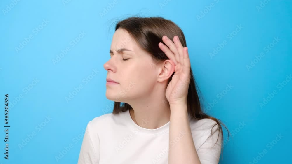 Portrait of confused young woman isolated on blue studio background having hearing problems, asking to say louder, difficult to listen quiet talk, wears t-shirt, misunderstanding in communication Portrait of confused young woman isolated on blue studio background having hearing problems, asking to say louder, difficult to listen quiet talk, wears t-shirt, misunderstanding in communication