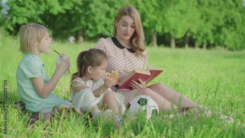 Portrait of attractive young mother reading book to children on green summer meadow. Happy Caucasian woman resting with son and daughter outdoors on sunny day. Leisure, family, lifestyle. Portrait of attractive young mother reading book to children on green summer meadow. Happy Caucasian woman resting with son and daughter outdoors on sunny day. Leisure, family, lifestyle.
