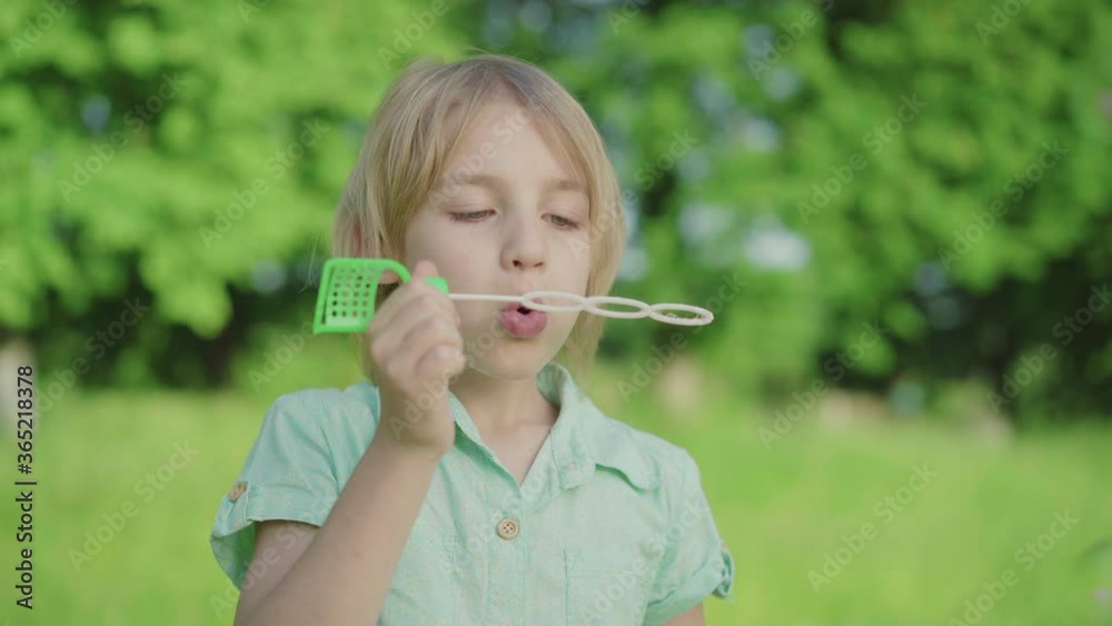 Close-up of little Caucasian boy blowing soap bubbles on sunny meadow. Cute blond child with green eyes resting outdoors on summer day. Leisure, joy, childhood, relaxation. Close-up of little Caucasian boy blowing soap bubbles on sunny meadow. Cute blond child with green eyes resting outdoors on summer day. Leisure, joy, childhood, relaxation.