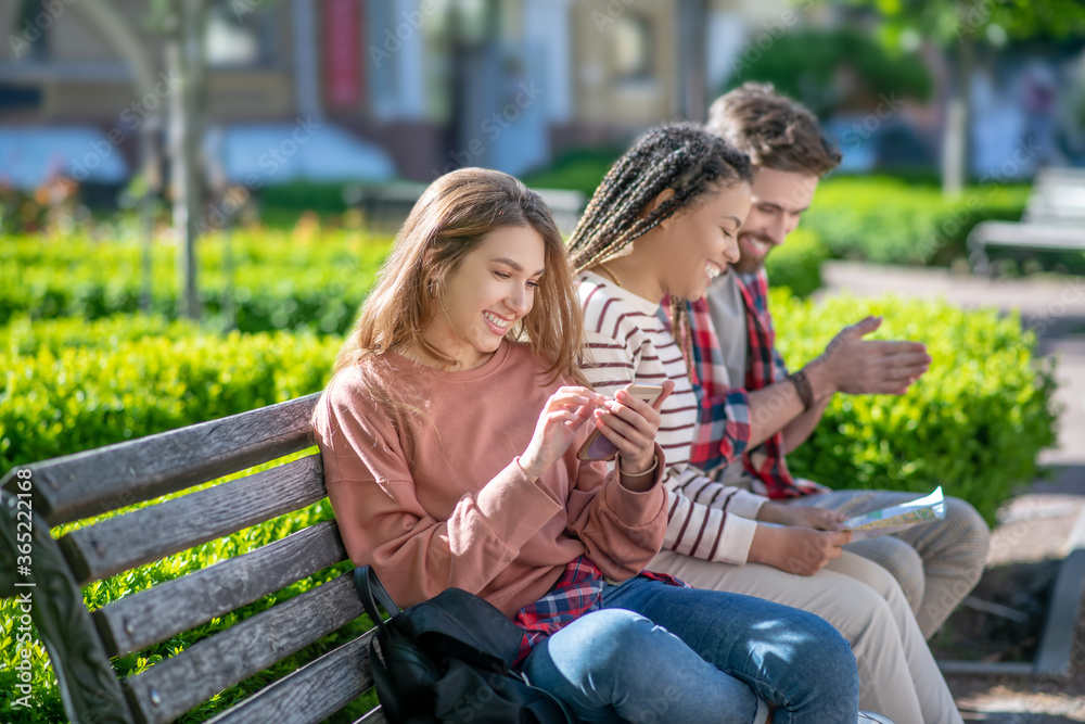 Girl with smartphone and girlfriend with guy with map on bench
