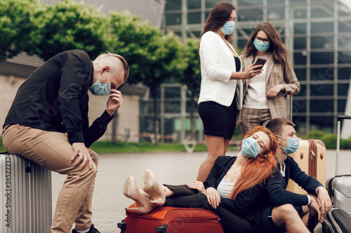 Close up portrait of a five friends waiting for the flight, sitting on the asphalt, using mobile phones. Three girls and two boys in protective masks with suitcases go on a trip