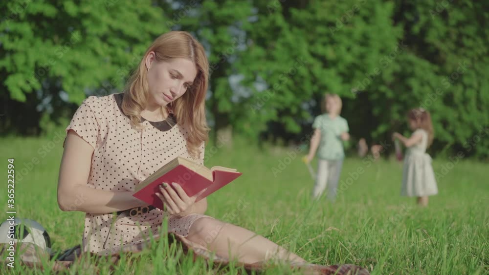 Positive beautiful mother reading book on green meadow and looking back at children playing at the background. Young Caucasian woman resting with son and daughter in park on weekends.