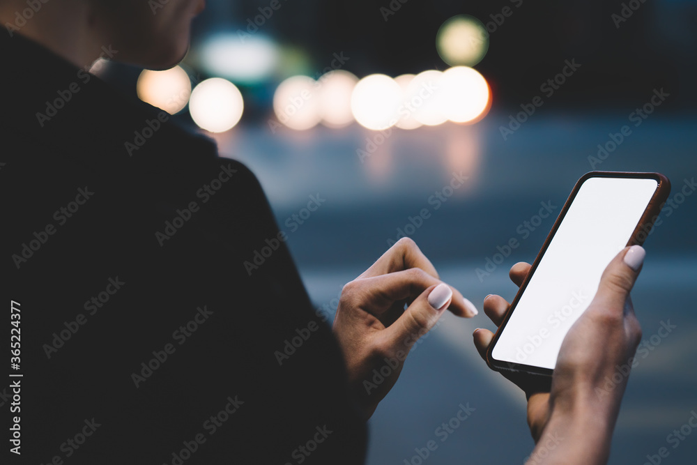 Cropped side view of woman's hands holding smartphone device with blank screen area for your website and installing application using 4G internet on bokeh background with night lights - obrazy, fototapety, plakaty 