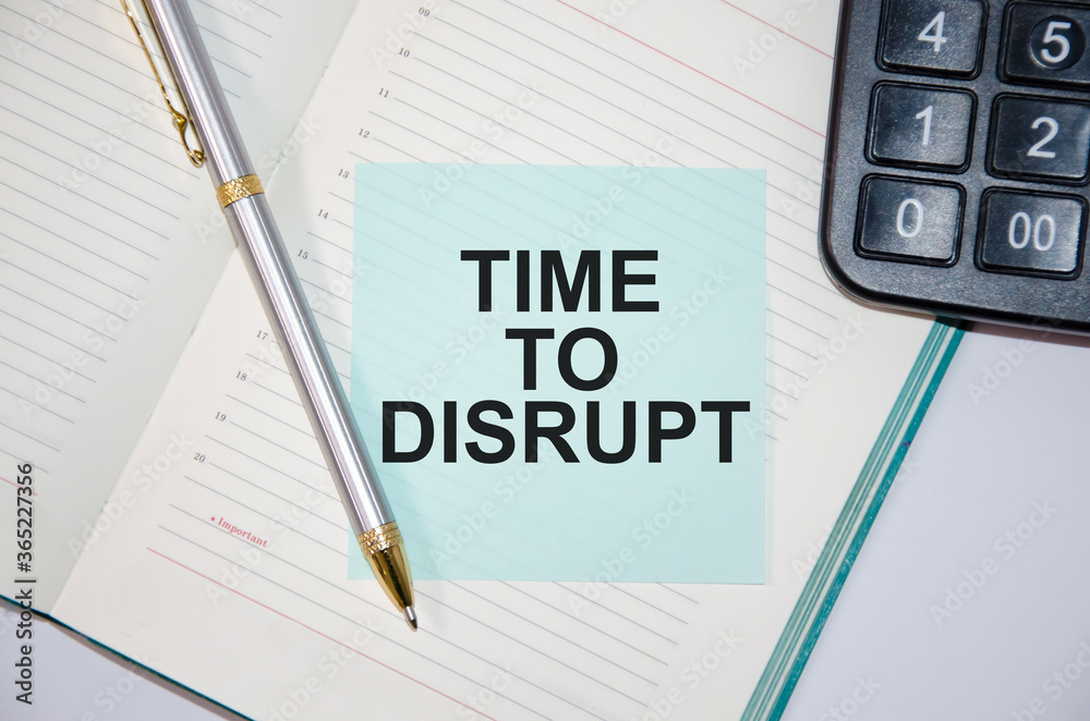 The word time to disrupt is written on a paper blue block near a calculator and notepad. pen next The word time to disrupt is written on a paper blue block near a calculator and notepad. pen next