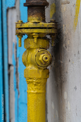 Old yellow gas valve, flange and screws on vertical pipe near wall. Valve is part of gas supply system of building, provide gas shutdown. Valve some rusty, covered spider web. Vertical, copy space.