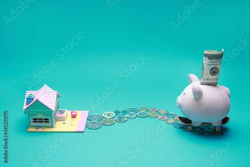 Small toy pink piggy bank with one hundred dollars on skateboard moving on coins road to toy house isolated on green background with copy space