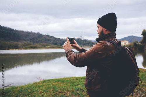 Young hipster guy taking photo on his mobile phone camera of a beautiful landscape while standing near lake, bearded man shoots video on cell telephone during amazing travel through the countryside