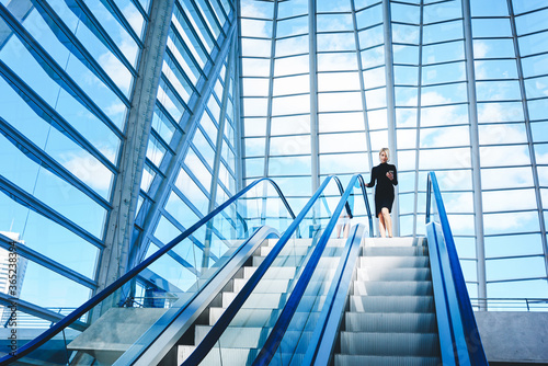 Female skilled CEO is dialing a number on mobile phone while is standing near escalator in interior with contemporary design. Woman is using cell telephone, while is moving on staircase in office