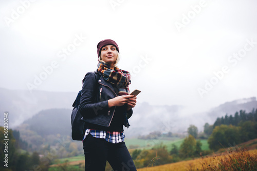 Stylish female tourist holding cell telephone while enjoy the beautiful landscape of mountains,trendy woman traveler orienteering on compass with mobile phone apps while hiking with backpack in forest