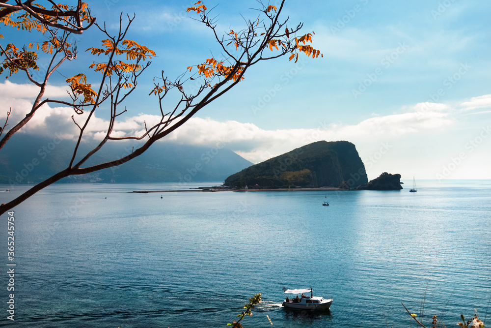 Blue sea water, lonely island and cumulus clouds at sunrise. White clouds sky reflections in water. Red leaves branch and boats at foreground