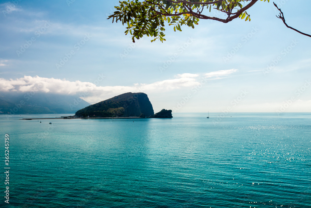 Aqua green sea water, lonely island and cumulus clouds at sunrise. White clouds sky reflections in water. Green leaves branch at foreground