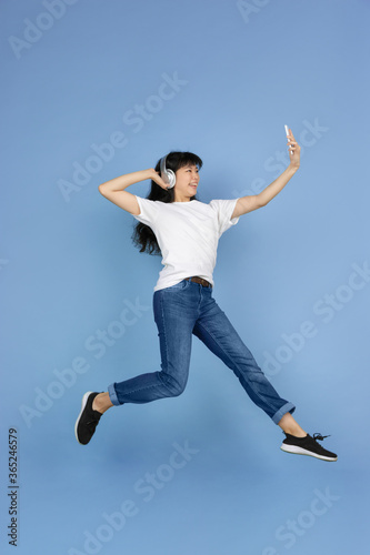 Jumping high, taking selfie with headphones. Portrait of young asian woman isolated on blue background. Beautiful cute girl. Human emotions, facial expression, sales, ad, online shopping concept.