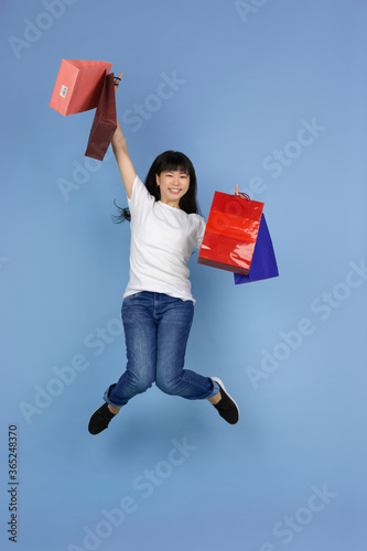 Jumping high with shopping bags, in flight. Portrait of young asian woman isolated on blue background. Beautiful cute girl. Human emotions, facial expression, sales, ad, online shopping concept.