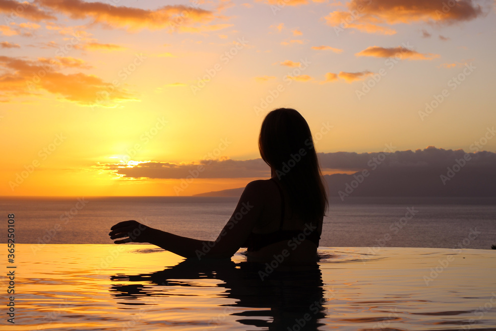  A girl in a swimsuit with her hair down stands with her back, her hand on the edge of the pool and looks at the sunset and the ocean.