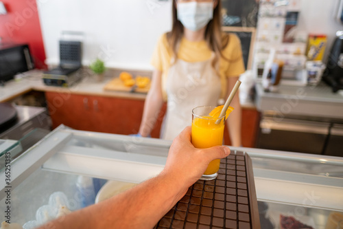 Woman at work in a bar wearing protective face mask - Hand of man who takes the fresh made orange juice from the counter - Workplaces follow the safety measures - New normality - Focus on the juice