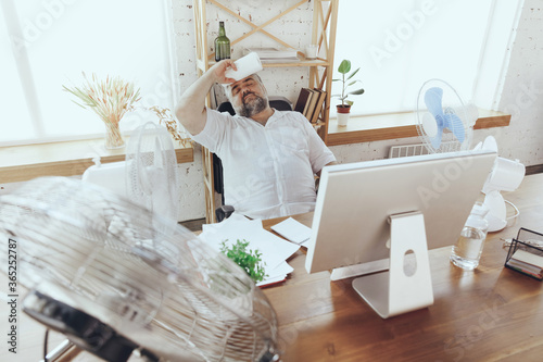 Wind. Businessman, manager in office with computer and fan cooling off, feeling hot, flushed. Using fan but still suffering of uncomfortable climate in cabinet. Summer, office working, business.