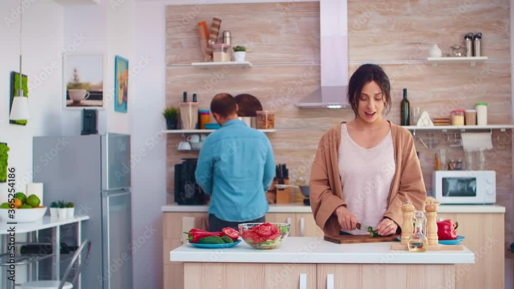 Woman slicing cucumbers on wooden cutting board for healthy salad. Husband opening refrigerator. Cooking preparing healthy organic food happy together lifestyle. Cheerful meal in family with Woman slicing cucumbers on wooden cutting board for healthy salad. Husband opening refrigerator. Cooking preparing healthy organic food happy together lifestyle. Cheerful meal in family with
