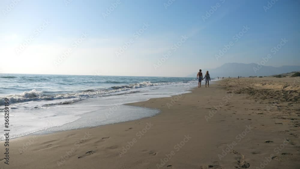 Couple of lovers walking at seashore holding hands during resting on resort. Man and woman strolling on coast enjoying summer vacation or their honeymoon. View on beach with seascape at background Couple of lovers walking at seashore holding hands during resting on resort. Man and woman strolling on coast enjoying summer vacation or their honeymoon. View on beach with seascape at background