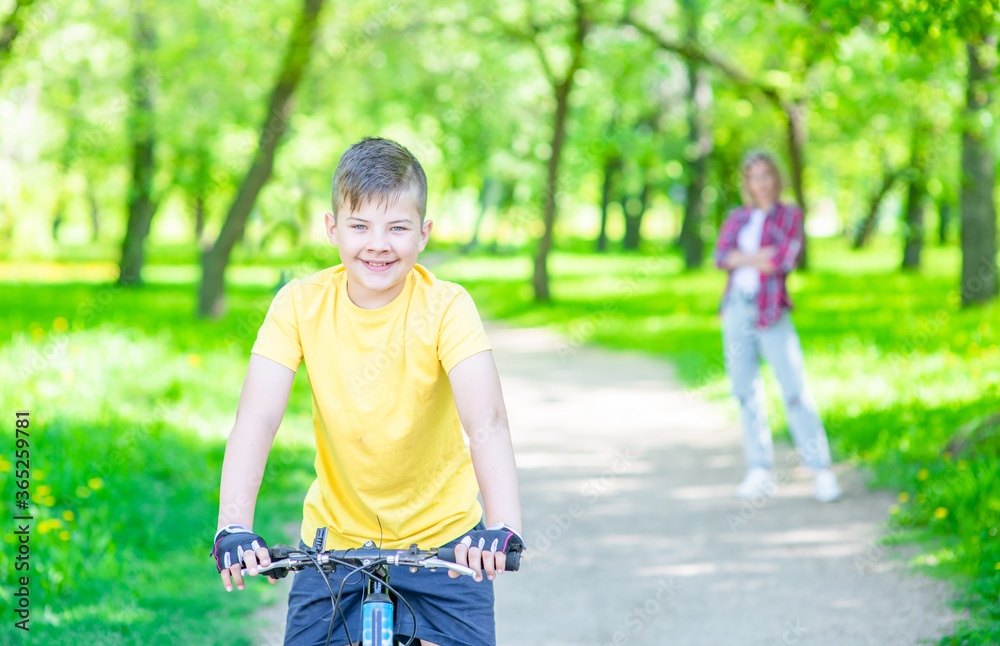 Smiling boy riding a bicycle in summer sunny park. Mom stands on the background Smiling boy riding a bicycle in summer sunny park. Mom stands on the background