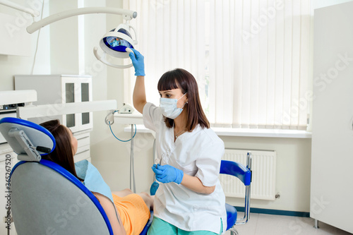 Young woman dentist with mask is adjusting the light in dentistry and prepare to examine the patient teeth