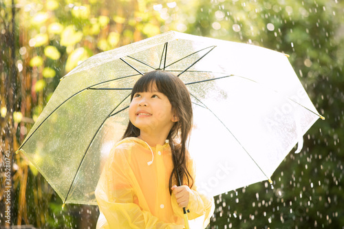 Asian girl is wearing yellow raincoat and enjoying rainfall in the park. Kid playing on the nature outdoors.
