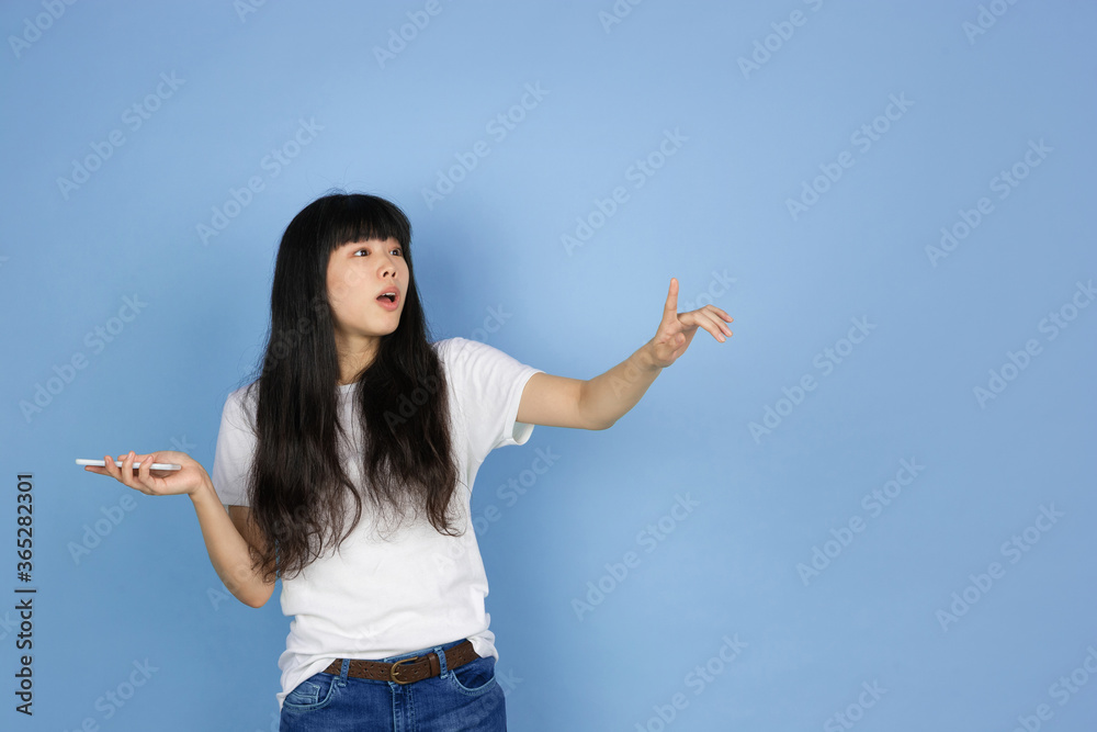 Using phone, pointing, shocked. Portrait of young asian woman isolated on blue studio background. Beautiful cute girl in casual. Human emotions, facial expression, sales, ad, online shopping concept. Using phone, pointing, shocked. Portrait of young asian woman isolated on blue studio background. Beautiful cute girl in casual. Human emotions, facial expression, sales, ad, online shopping concept.