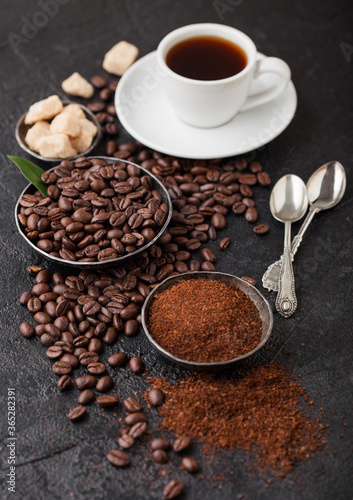 White cup of fresh raw organic coffee with beans and ground powder with cane sugar cubes with coffee tree leaf on black background. Top view. Silver tea spoons